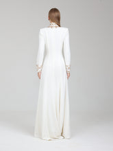 Load image into Gallery viewer, A woman viewed from the back wearing a long white or ivory floor-length gown with long sleeves and ornate gold detailing at the high neckline and cuffs, photographed against a light gray studio background.