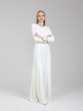 Load image into Gallery viewer, A woman modeling a long white floor-length gown with a high mock neck, long sleeves, and ornate gold detailing around the collar and cuffs, standing against a light gray studio background.