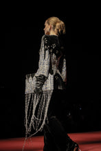 Load image into Gallery viewer, A model walks a fashion runway wearing a black velvet jacket with intricate silver beadwork, sequins, and a long fringe detail, paired with black pants against a dark stage background.