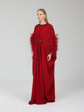 Load image into Gallery viewer, A woman modeling a floor-length red gown with wide draped sleeves, feathered trim on the sleeves, a beaded neckline, and a beaded waistband, photographed against a light gray studio background.
