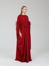 Load image into Gallery viewer, A woman modeling a floor-length red gown with feathered cape sleeves and a dark embroidered waistband, photographed against a plain light gray studio background.
