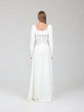 Load image into Gallery viewer, A woman viewed from the back wearing a floor-length white gown with long sleeves, a square neckline, and ornate beaded embellishment across the upper back and along the sleeve cuffs, photographed against a light gray studio background.

